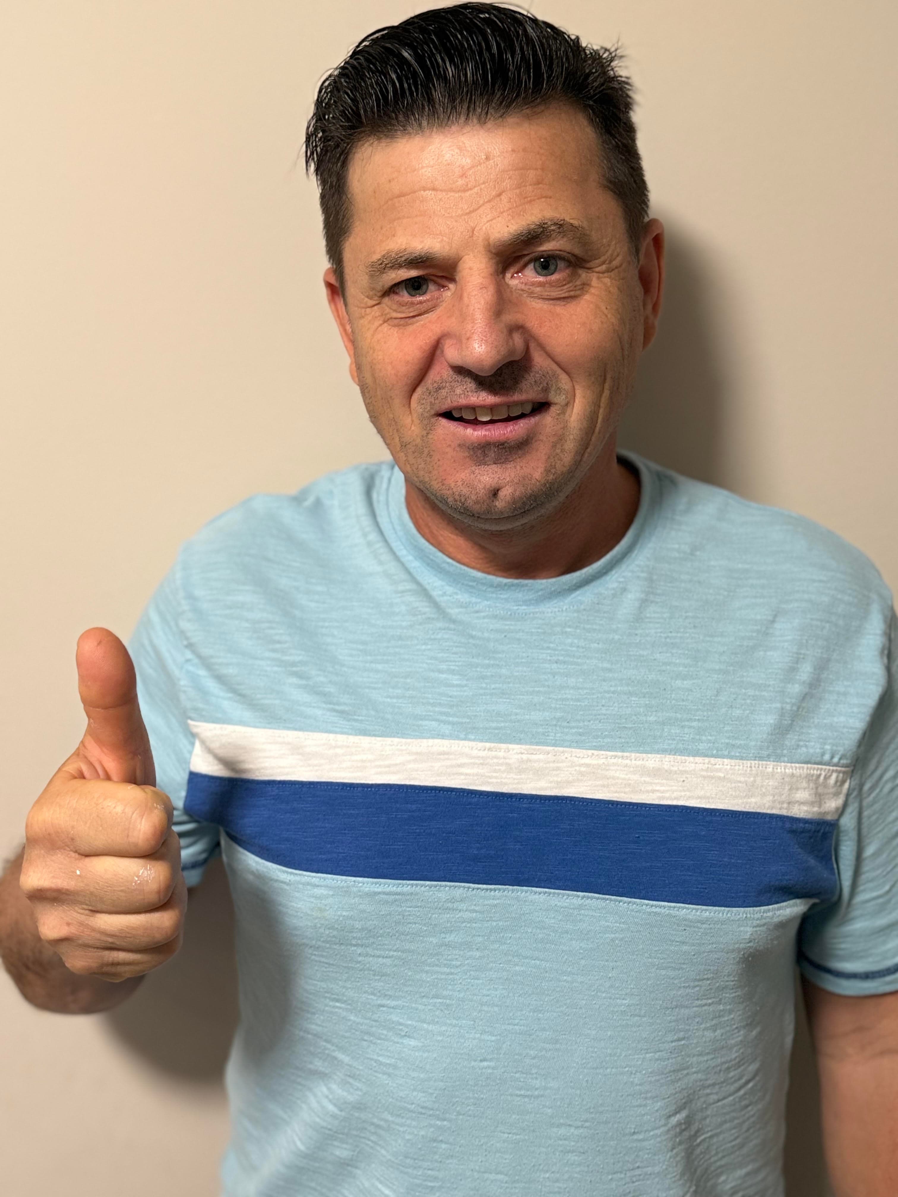 Smiling man in a blue striped t-shirt giving a thumbs-up gesture against a neutral background.